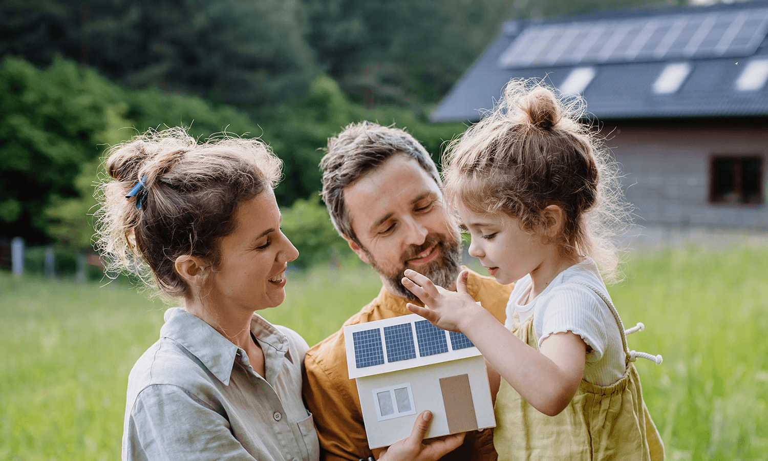Familie mit einem Modellhaus auf einem großen Grundstück mit Wald und echtem Haus im Hintergrund.